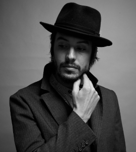 Black and white thumbnail of American blues singer Cory Chisel wearing a Homburg style hat and stripe, patterned tweed, woollen overcoat with upturned collar