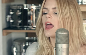 Co;our Image of versatile blonde, Dutch vocalist Davina Michelle singing into a silver microphone in a music studio with out0of-focus drums on unvarnished wooden shelves behind her